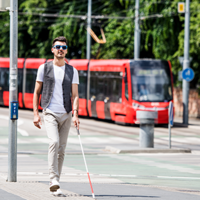 Ein junger blinder Mann läuft auf dem Gehweg. Er hat eine Sonnenbrille auf und einen Blindenstock in der Hand. Mit dem Blindenstock benutzt er die Leitstreifen auf dem Boden. Im Hintergrund fährt eine Straßenbahn entlang.