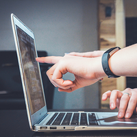 Zwei Personen arbeiten gemeinsam an einem Laptop. Eine Person zeigt mit dem Finger auf den Bildschirm, während die andere Person die Tastatur bedient. Beide Hände sind im Vordergrund zu sehen, und eine der Personen trägt ein Armband. Der Hintergrund ist unscharf, wodurch der Fokus auf den Laptop und die Hände gelenkt wird.