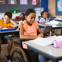 In einem Klassenzimmer sitzen mehrere Kinder an ihren Tischen und arbeiten konzentriert. Im Vordergrund sitzt ein Mädchen im Rollstuhl, das ein Tablet in den Händen hält und lächelt. Auf ihrem Tisch liegen Schulmaterialien wie Hefte und ein Federmäppchen. 