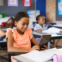 In einem Klassenzimmer sitzen mehrere Kinder an ihren Tischen und arbeiten konzentriert. Im Vordergrund sitzt ein Mädchen im Rollstuhl, das ein Tablet in den Händen hält und lächelt. Auf ihrem Tisch liegen Schulmaterialien wie Hefte und ein Federmäppchen. 