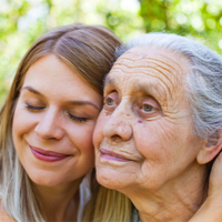 Auf dem Bild sind zwei Frauen zu sehen, die sich liebevoll aneinander schmiegen. Die jüngere Frau hat lange, blonde Haare und lächelt mit geschlossenen Augen. Die ältere Frau hat graue Haare und schaut in die Ferne. Beide befinden sich im Freien vor einem Hintergrund aus unscharfem Grün, das auf Bäume oder Pflanzen hinweist. 