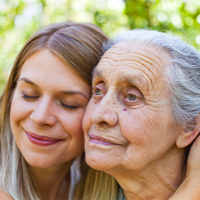 Auf dem Bild sind zwei Frauen zu sehen, die sich liebevoll aneinander schmiegen. Die jüngere Frau hat lange, blonde Haare und lächelt mit geschlossenen Augen. Die ältere Frau hat graue Haare und schaut in die Ferne. Beide befinden sich im Freien vor einem Hintergrund aus unscharfem Grün, das auf Bäume oder Pflanzen hinweist. 