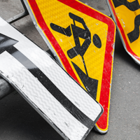 Auf dem Bild sind mehrere Verkehrsschilder zu sehen, die auf dem Boden liegen. Eines der Schilder zeigt einen schwarzen Pfeil, der nach links weist, auf weißem Hintergrund. Ein anderes Schild zeigt ein Symbol für Straßenbauarbeiten: eine stilisierte Figur, die mit einer Schaufel arbeitet, auf gelbem Hintergrund mit rotem Rand. Ein weiteres Schild zeigt eine kurvige Straße, ebenfalls auf gelbem Hintergrund mit rotem Rand.