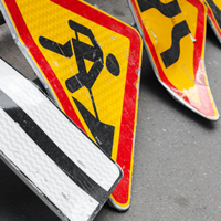 Auf dem Bild sind mehrere Verkehrsschilder zu sehen, die auf dem Boden liegen. Eines der Schilder zeigt einen schwarzen Pfeil, der nach links weist, auf weißem Hintergrund. Ein anderes Schild zeigt ein Symbol für Straßenbauarbeiten: eine stilisierte Figur, die mit einer Schaufel arbeitet, auf gelbem Hintergrund mit rotem Rand. Ein weiteres Schild zeigt eine kurvige Straße, ebenfalls auf gelbem Hintergrund mit rotem Rand.