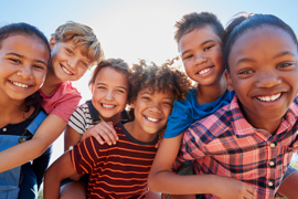 Eine Gruppe von sechs fröhlichen Kindern steht dicht beieinander und lächelt in die Kamera. Sie sind bunt gekleidet. 