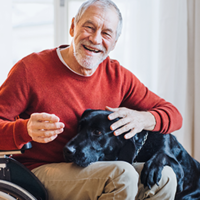 Ein älterer Mann sitzt in einem Rollstuhl und trägt einen roten Pullover sowie beige Hosen. Er lächelt herzlich und streichelt einen großen schwarzen Hund, der seinen Kopf auf den Schoß des Mannes gelegt hat. Im Hintergrund sind helle Vorhänge und ein Fenster zu sehen, durch das Tageslicht hereinfällt. 