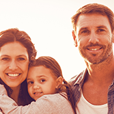 Auf dem Bild ist eine Familie. Links steht die Frau, hat das Mädchen auf dem Arm. Rechts steht ein Mann, der Frau mit Kind zugewandt. Im Hintergrund steht links neben der Familie eine alte Frau und ein alter Mann.