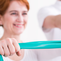 Eine Frau führt eine Übung mit einem grünen Fitnessband durch. Die Frau lächelt, während sie das Band mit beiden Händen hält und es auseinanderzieht. Sie trägt helle Kleidung, und der Hintergrund ist unscharf.