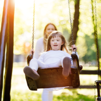 Ein Kind schaukelt auf einer Schaukel im Park. Im Hintergrund ist eine erwachsene Person zu sehen, die das Kind anschubst. Die Szene ist von hellem Sonnenlicht durchflutet, das durch die Bäume im Hintergrund scheint. Das Kind trägt helle Kleidung. 