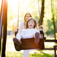 Ein Kind schaukelt auf einer Schaukel im Park. Im Hintergrund ist eine erwachsene Person zu sehen, die das Kind anschubst. Die Szene ist von hellem Sonnenlicht durchflutet, das durch die Bäume im Hintergrund scheint. Das Kind trägt helle Kleidung. 