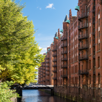 Das Foto ist von einer Brücke mit Blick auf einen Kanal in der Speicherstadt ausgenommen. Auf der linken Seite stehen hellgrüne Bäume und auf der rechten Seite sind hohe Gebäude mit Rot-Klinker-Fassade.