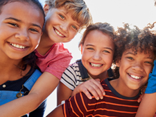 Eine Gruppe von sechs fröhlichen Kindern steht dicht beieinander und lächelt in die Kamera. Sie sind bunt gekleidet.