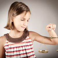 Ein junges Mädchen mit schulterlangem braunem Haar hält eine goldene Waage in ihrer rechten Hand. Sie trägt ein ärmelloses Oberteil mit Streifen in verschiedenen Farben und schaut konzentriert auf die Waage. Der Hintergrund ist neutral und grau.