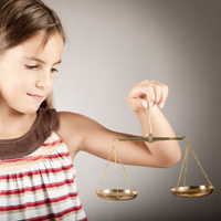 Ein junges Mädchen mit schulterlangem braunem Haar hält eine goldene Waage in ihrer rechten Hand. Sie trägt ein ärmelloses Oberteil mit Streifen in verschiedenen Farben und schaut konzentriert auf die Waage. Der Hintergrund ist neutral und grau.
