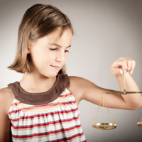 Ein junges Mädchen mit schulterlangem braunem Haar hält eine goldene Waage in ihrer rechten Hand. Sie trägt ein ärmelloses Oberteil mit Streifen in verschiedenen Farben und schaut konzentriert auf die Waage. Der Hintergrund ist neutral und grau.