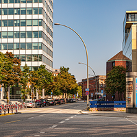 Eine breite, mehrspurige Straße verläuft durch ein urbanes Gebiet mit modernen Bürogebäuden und Bäumen am Straßenrand. Links ist ein Eingang zur U-Bahn-Station erkennbar. Mehrere geparkte Autos stehen am Straßenrand, daneben befinden sich Fahrradständer mit Leihfahrrädern. Rechts ragt ein rotes Backsteingebäude mit Glasfassade in das Bild. Die Straße ist fast leer, der Himmel ist blau und klar. 