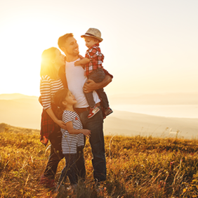 Eine Familie steht auf einer Wiese bei Sonnenuntergang. Ein Mann hält ein kleines Kind im Arm, während eine Frau und ein weiteres Kind neben ihnen stehen. Alle lächeln. Im Hintergrund erstreckt sich eine Landschaft mit Hügeln und einem Gewässer.