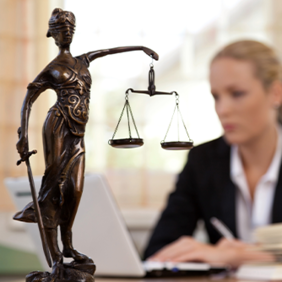 Das Bild zeigt eine Frau in einem Büro, die an einem Laptop arbeitet. Im Vordergrund steht eine Statue der Justitia, die Göttin der Gerechtigkeit, mit einer Waage in der einen und einem Schwert in der anderen Hand. Die Frau im Hintergrund ist unscharf dargestellt, während die Statue scharf fokussiert ist. Auf dem Schreibtisch vor ihr liegt ein aufgeschlagenes Buch.