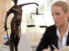 Das Bild zeigt eine Frau in einem Büro, die an einem Laptop arbeitet. Im Vordergrund steht eine Statue der Justitia, die Göttin der Gerechtigkeit, mit einer Waage in der einen und einem Schwert in der anderen Hand. Die Frau im Hintergrund ist unscharf dargestellt, während die Statue scharf fokussiert ist. Auf dem Schreibtisch vor ihr liegt ein aufgeschlagenes Buch. 