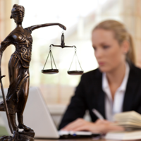Das Bild zeigt eine Frau in einem Büro, die an einem Laptop arbeitet. Im Vordergrund steht eine Statue der Justitia, die Göttin der Gerechtigkeit, mit einer Waage in der einen und einem Schwert in der anderen Hand. Die Frau im Hintergrund ist unscharf dargestellt, während die Statue scharf fokussiert ist. Auf dem Schreibtisch vor ihr liegt ein aufgeschlagenes Buch. 
