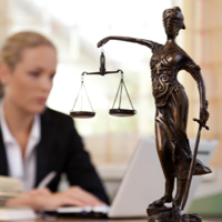 Das Bild zeigt eine Frau in einem Büro, die an einem Laptop arbeitet. Im Vordergrund steht eine Statue der Justitia, die Göttin der Gerechtigkeit, mit einer Waage in der einen und einem Schwert in der anderen Hand. Die Frau im Hintergrund ist unscharf dargestellt, während die Statue scharf fokussiert ist. Auf dem Schreibtisch vor ihr liegt ein aufgeschlagenes Buch. 