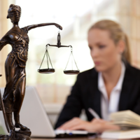 Das Bild zeigt eine Frau in einem Büro, die an einem Laptop arbeitet. Im Vordergrund steht eine Statue der Justitia, die Göttin der Gerechtigkeit, mit einer Waage in der einen und einem Schwert in der anderen Hand. Die Frau im Hintergrund ist unscharf dargestellt, während die Statue scharf fokussiert ist. Auf dem Schreibtisch vor ihr liegt ein aufgeschlagenes Buch. 