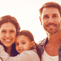 Auf dem Bild ist eine Familie. Links steht die Frau, hat das Mädchen auf dem Arm. Rechts steht ein Mann, der Frau mit Kind zugewandt.