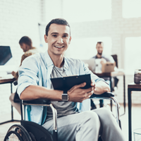 Ein Mann sitzt in einem Rollstuhl und hält ein Tablet in den Händen. Er lächelt und trägt ein hellblaues Hemd über einem grauen T-Shirt. Im Hintergrund befinden sich mehrere Personen, die an Schreibtischen arbeiten.