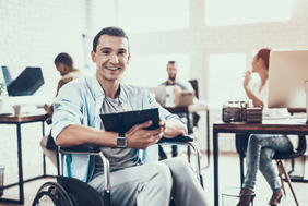 Ein Mann sitzt in einem Rollstuhl und hält ein Tablet in den Händen. Er lächelt und trägt ein hellblaues Hemd über einem grauen T-Shirt. Im Hintergrund befinden sich mehrere Personen, die an Schreibtischen arbeiten.