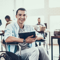 Ein Mann sitzt in einem Rollstuhl und hält ein Tablet in den Händen. Er lächelt und trägt ein hellblaues Hemd über einem grauen T-Shirt. Im Hintergrund befinden sich mehrere Personen, die an Schreibtischen arbeiten.
