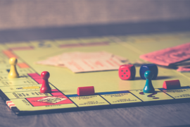 Ein klassisches Monopoly-Spielbrett liegt auf einem Tisch. Mehrere bunte Spielfiguren, rote Häuser, zwei Würfel und Spielgeld sind auf dem Brett verteilt. Die Spielfiguren befinden sich auf verschiedenen Feldern, unter anderem auf einem Eckfeld. Im Hintergrund liegen Karten und Spielgeld unscharf auf dem Brett. 