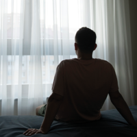 Ein Mann sitzt auf einem Bett und schaut aus einem großen Fenster mit weißen Vorhängen. Das Zimmer ist in sanftes Licht getaucht, das durch die Vorhänge fällt. Im Hintergrund sind unscharf Gebäude zu erkennen. Der Mann trägt ein T-Shirt und hat kurze Haare.