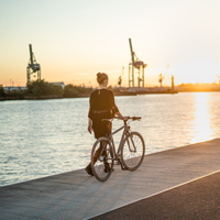 Auf dem Bild steht eine Frau mit ihrem Fahrrad an einer Kaimauer an einem Fluss. Im Hintergrund ist ein Fluss und ein Hafen zu sehen. Die Sonne geht unter.