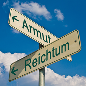 Das Bild zeigt zwei Straßenschilder, die in entgegengesetzte Richtungen zeigen. Auf dem Schild, das nach links weist, steht das Wort "Armut" und auf dem Schild, das nach rechts weist, steht das Wort "Reichtum". Der Himmel im Hintergrund ist blau mit einigen weißen Wolken.
