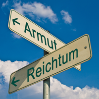 Das Bild zeigt zwei Straßenschilder, die in entgegengesetzte Richtungen zeigen. Auf dem Schild, das nach links weist, steht das Wort "Armut" und auf dem Schild, das nach rechts weist, steht das Wort "Reichtum". Der Himmel im Hintergrund ist blau mit einigen weißen Wolken.