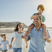 Eine Familie läuft lachend am Strand entlang. Ein Mann trägt ein Kind auf den Schultern, neben ihm gehen eine Frau und ein weiteres Kind. Im Hintergrund sind Meer, Wellen und ein Hügel zu sehen, die Sonne scheint.