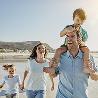Eine Familie läuft lachend am Strand entlang. Ein Mann trägt ein Kind auf den Schultern, neben ihm gehen eine Frau und ein weiteres Kind. Im Hintergrund sind Meer, Wellen und ein Hügel zu sehen, die Sonne scheint.