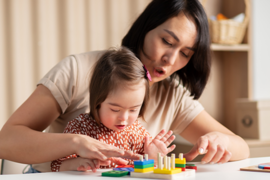 Eine Frau und ein kleines Mädchen sitzen an einem Tisch und spielen mit bunten Holzspielzeugen. Die Frau zeigt dem Mädchen, wie man die Spielzeuge benutzt, während das Mädchen konzentriert zuschaut und mit den Händen die Teile berührt. 