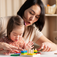 Eine Frau und ein kleines Mädchen sitzen an einem Tisch und spielen mit bunten Holzspielzeugen. Die Frau zeigt dem Mädchen, wie man die Spielzeuge benutzt, während das Mädchen konzentriert zuschaut und mit den Händen die Teile berührt. 
