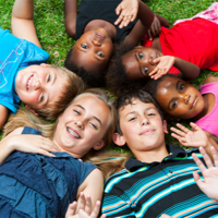 Auf dem Bild liegen sechs Kinder auf dem Rasen in einem Kreis und lächeln in die Kamera. Sie haben ihre Köpfe nah beieinander, sodass sie einen geschlossenen Kreis bilden. Die Kinder tragen bunte Sommerkleidung und scheinen fröhlich und entspannt zu sein. Einige von ihnen winken der Kamera zu. Der Hintergrund besteht aus grünem Gras.