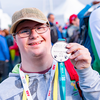 Ein junger Mann mit Brille und Baseballkappe lächelt in die Kamera und hält stolz eine Medaille hoch. Er trägt eine graue Kapuzenjacke und hat ein buntes Band um den Hals. Im Hintergrund sind weitere Personen zu sehen, die sich im Freien aufhalten. Die Atmosphäre wirkt fröhlich und feierlich.