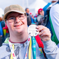 Ein junger Mann mit Brille und Baseballkappe lächelt in die Kamera und hält stolz eine Medaille hoch. Er trägt eine graue Kapuzenjacke und hat ein buntes Band um den Hals. Im Hintergrund sind weitere Personen zu sehen, die sich im Freien aufhalten. Die Atmosphäre wirkt fröhlich und feierlich.