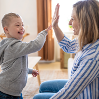 Ein kleiner Junge und eine Frau sitzen sich in einem gemütlichen Wohnzimmer gegenüber. Der Junge trägt einen grauen Kapuzenpullover und gibt der Frau, die ein gestreiftes Hemd trägt, ein High-Five. 