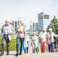 Eine Gruppe älterer Menschen geht eine Treppe hinauf. Sie tragen bunte Einkaufstaschen. Im Hintergrund sind moderne Bürogebäude und grüne Pflanzen zu sehen. Die Sonne scheint. Die Personen unterstützen sich gegenseitig beim Gehen.
