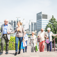 Eine Gruppe älterer Menschen geht eine Treppe hinauf. Sie tragen bunte Einkaufstaschen. Im Hintergrund sind moderne Bürogebäude und grüne Pflanzen zu sehen. Die Sonne scheint. Die Personen unterstützen sich gegenseitig beim Gehen.