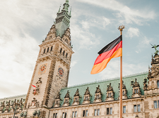 Das historische Rathaus in Hamburg mit seiner reich verzierten Fassade und dem hohen Uhrturm. Im Vordergrund weht eine deutsche Flagge.