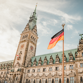 Das historische Rathaus in Hamburg mit seiner reich verzierten Fassade und dem hohen Uhrturm. Im Vordergrund weht eine deutsche Flagge.