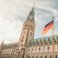 Das historische Rathaus in Hamburg mit seiner reich verzierten Fassade und dem hohen Uhrturm. Im Vordergrund weht eine deutsche Flagge.