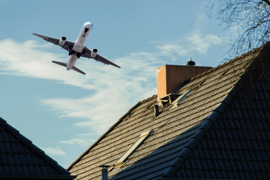 Ein Flugzeug fliegt knapp über die Dächer von Wohnhäusern. Der Himmel ist klar mit einigen Wolken, und die Dächer sind mit dunklen Ziegeln gedeckt. Ein Schornstein ist auf einem der Dächer zu sehen. 