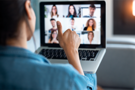 Eine Person sitzt vor einem Laptop und nimmt an einer Videokonferenz teil. Auf dem Bildschirm sind mehrere Teilnehmer in kleinen Fenstern zu sehen. Die Person im Vordergrund zeigt einen Daumen hoch, was auf eine positive Interaktion oder Zustimmung hinweisen könnte. Der Hintergrund ist unscharf, wodurch der Fokus auf die Handgeste und den Laptop gelegt wird.